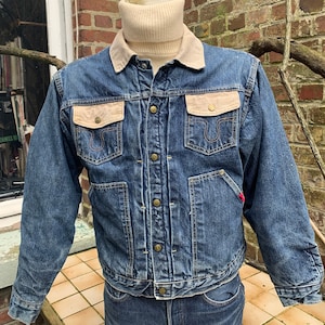 May include: A blue denim jacket with a brown corduroy collar and two large patch pockets on the chest. The jacket has a button closure and a red label on the bottom left side.
