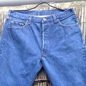 May include: A pair of medium-wash blue denim jeans featuring a button-fly closure, five pockets, and a straight-leg cut. The jeans are displayed on a black hanger, showing a classic construction with visible stitching and rolled-up leg cuffs.