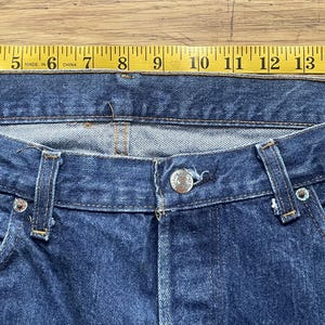 May include: Blue denim jeans featuring a metal button closure, belt loops, and a classic five-pocket design. A yellow measuring tape is positioned along the waistband, indicating a width of approximately 18 inches across the top edge.