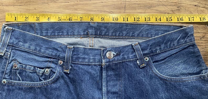 May include: Blue denim jeans featuring a metal button closure, belt loops, and a classic five-pocket design. A yellow measuring tape is positioned along the waistband, indicating a width of approximately 18 inches across the top edge.