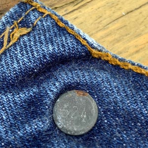 May include: A blue denim fabric section featuring a circular metal button embossed with the number 524. The denim is constructed with thick, contrasting orange stitching along the top edge, showcasing the texture of the woven cotton material.