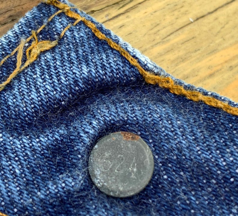 May include: A blue denim fabric section featuring a circular metal button embossed with the number 524. The denim is constructed with thick, contrasting orange stitching along the top edge, showcasing the texture of the woven cotton material.
