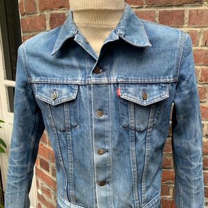 May include: A light blue denim jacket with a button closure and a red tab on the pocket. The jacket has a classic style and is in good condition.