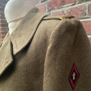 May include: A brown wool coat with a gold button on the shoulder and a red and black patch with a gold emblem on the sleeve.