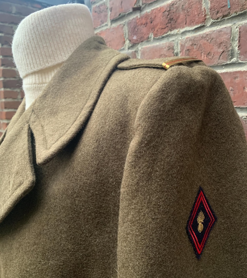 May include: A brown wool coat with a gold button on the shoulder and a red and black patch with a gold emblem on the sleeve.