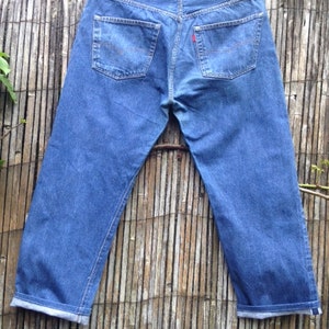 May include: A pair of blue denim jeans shown from the back, featuring two rear pockets with orange stitching, a tan leather patch on the waistband, and a small red tab on the right pocket. The pant legs are cuffed at the bottom hem.