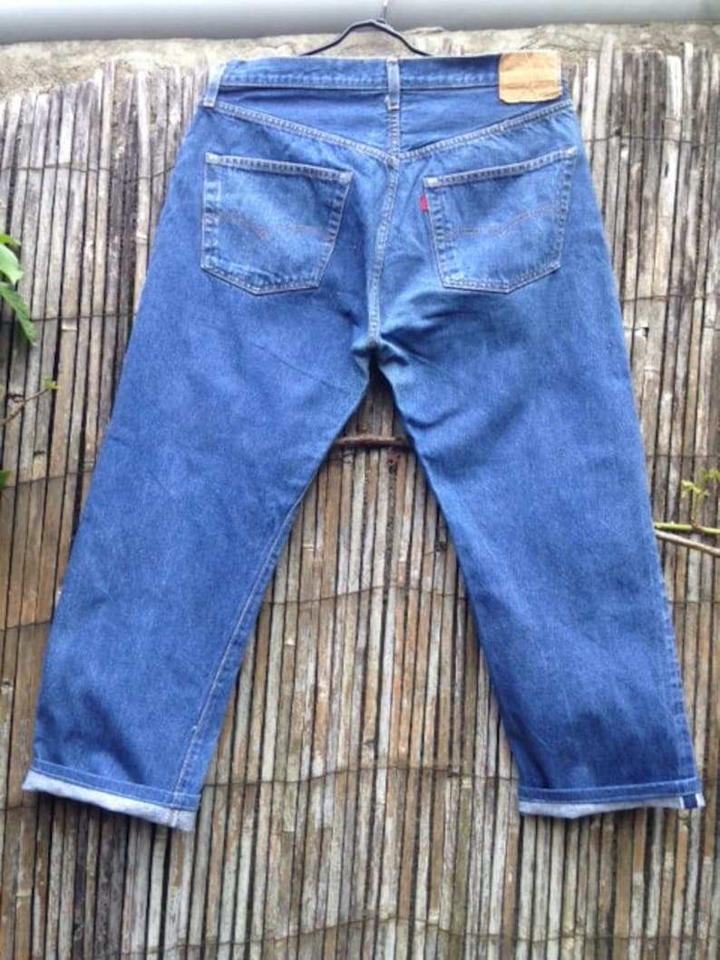 May include: A pair of blue denim jeans shown from the back, featuring two rear pockets with orange stitching, a tan leather patch on the waistband, and a small red tab on the right pocket. The pant legs are cuffed at the bottom hem.