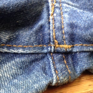 May include: A close-up view of blue denim fabric featuring a prominent horizontal seam and a vertical seam intersection. The denim has a textured, woven appearance with visible fading and thick, contrasting orange stitching along the seams.
