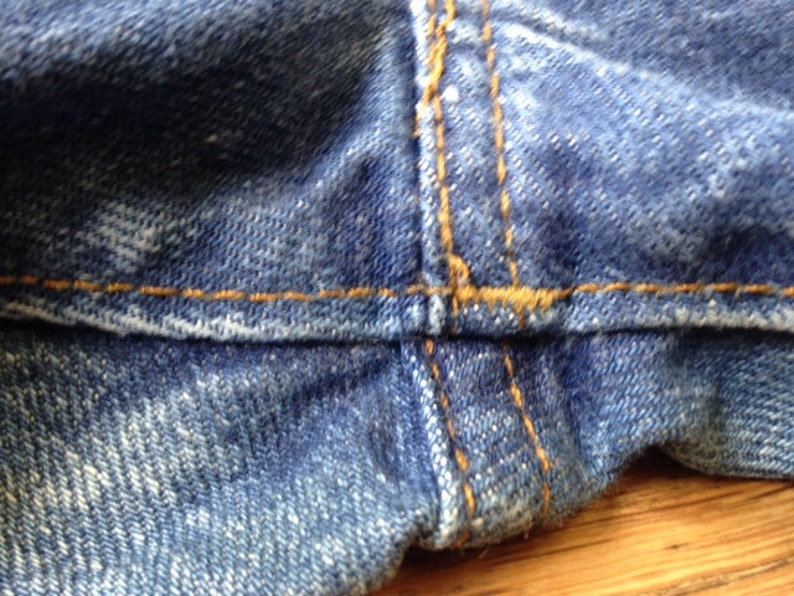 May include: A close-up view of blue denim fabric featuring a prominent horizontal seam and a vertical seam intersection. The denim has a textured, woven appearance with visible fading and thick, contrasting orange stitching along the seams.
