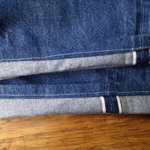 May include: A pair of dark blue denim jeans featuring a prominent selvedge edge on the cuffs. The rolled-up hems reveal the characteristic white and red stitching detail of the selvedge fabric, showcasing the heavy-duty construction of the denim.