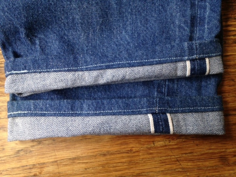May include: A pair of dark blue denim jeans featuring a prominent selvedge edge on the cuffs. The rolled-up hems reveal the characteristic white and red stitching detail of the selvedge fabric, showcasing the heavy-duty construction of the denim.