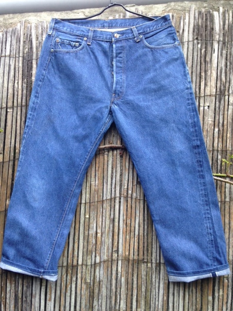 May include: A pair of medium-wash blue denim jeans featuring a button-fly closure, five pockets, and a straight-leg cut. The jeans are displayed on a black hanger, showing a classic construction with visible stitching and rolled-up leg cuffs.