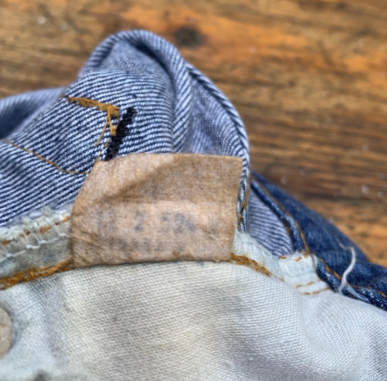 May include: A blue and white striped denim fabric featuring a tan rectangular interior garment tag with the printed text 12 2 53. The denim is constructed with visible orange stitching along the seams and a white interior lining fabric.