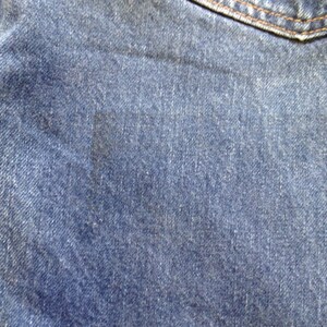 May include: A pair of blue denim jeans featuring a classic twill weave texture, a visible pocket seam at the top, and a distinct dark rectangular discoloration mark on the fabric surface, showcasing the durable cotton material and stitching.