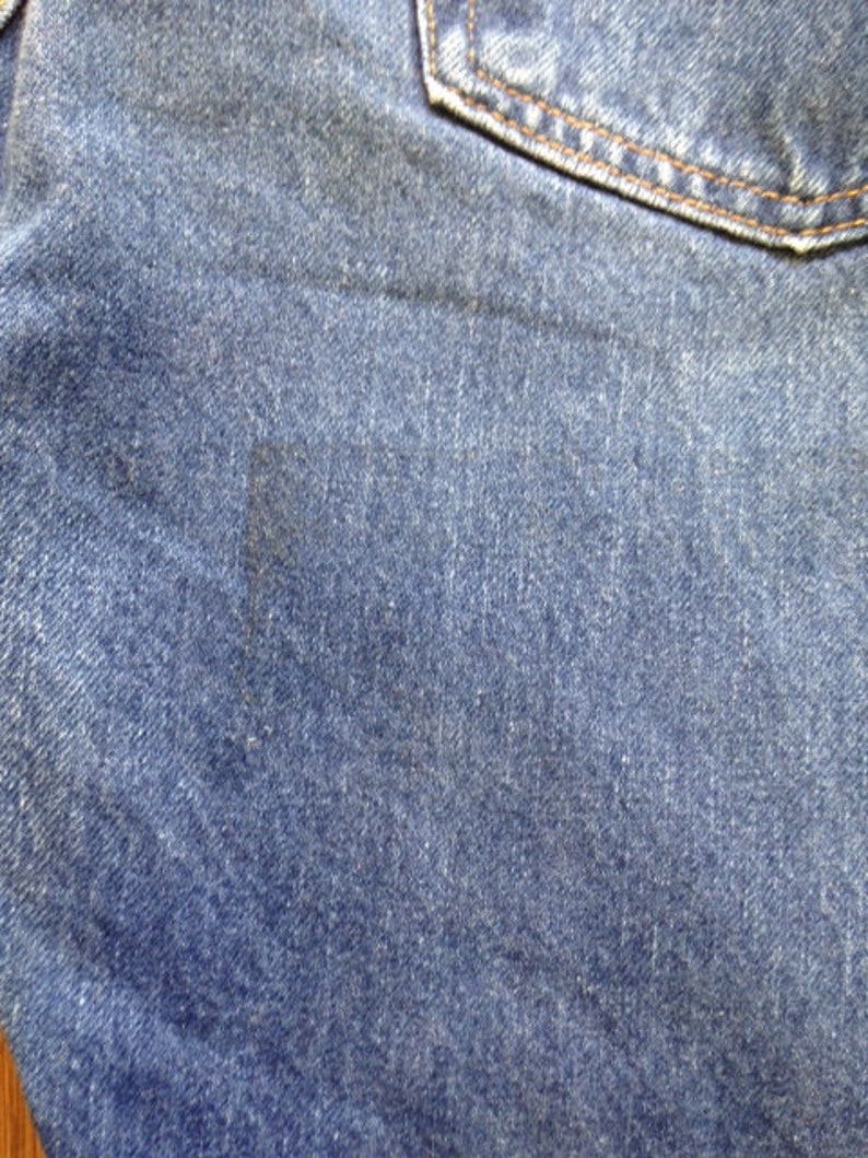 May include: A pair of blue denim jeans featuring a classic twill weave texture, a visible pocket seam at the top, and a distinct dark rectangular discoloration mark on the fabric surface, showcasing the durable cotton material and stitching.