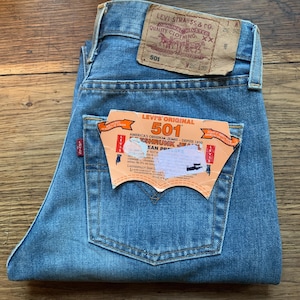 Peut inclure: Jean Levi's 501 bleu clair plié. La poche arrière porte une étiquette avec le logo Levi's et le texte "501". Un patch en cuir marron avec le logo Levi Strauss & Co. est visible à la taille. Le jean est de taille 26.