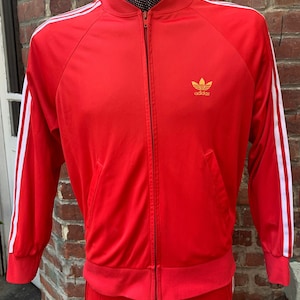 Peut inclure: Veste de survêtement Adidas rouge avec des bandes blanches et un logo doré. La veste a une fermeture éclair et un col montant.