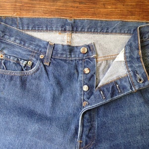 May include: A pair of dark blue denim jeans featuring a button-fly closure with four visible silver-toned metal buttons, a standard waistband with belt loops, a small coin pocket, and reinforced copper-colored rivets at the pocket corners.