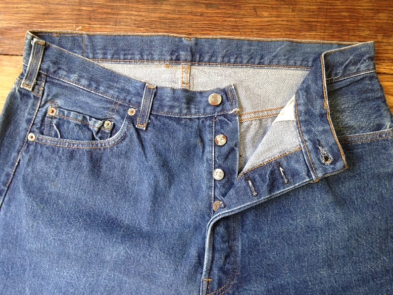 May include: A pair of dark blue denim jeans featuring a button-fly closure with four visible silver-toned metal buttons, a standard waistband with belt loops, a small coin pocket, and reinforced copper-colored rivets at the pocket corners.