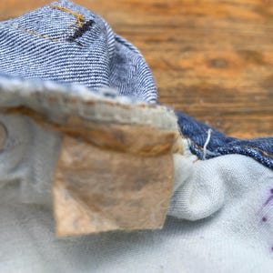 May include: A close-up view of the interior waistband of blue denim jeans, featuring a worn, light brown rectangular fabric label attached with stitching. The denim fabric shows a textured weave with visible orange thread details along the seams.