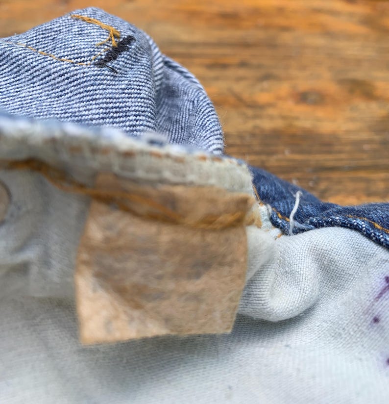 May include: A close-up view of the interior waistband of blue denim jeans, featuring a worn, light brown rectangular fabric label attached with stitching. The denim fabric shows a textured weave with visible orange thread details along the seams.