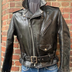 May include: A black leather motorcycle jacket featuring a wide notched collar, an asymmetrical silver-tone metal zip closure, multiple zipped pockets, shoulder epaulettes, and a buckled waist belt. The jacket is worn over blue denim jeans.