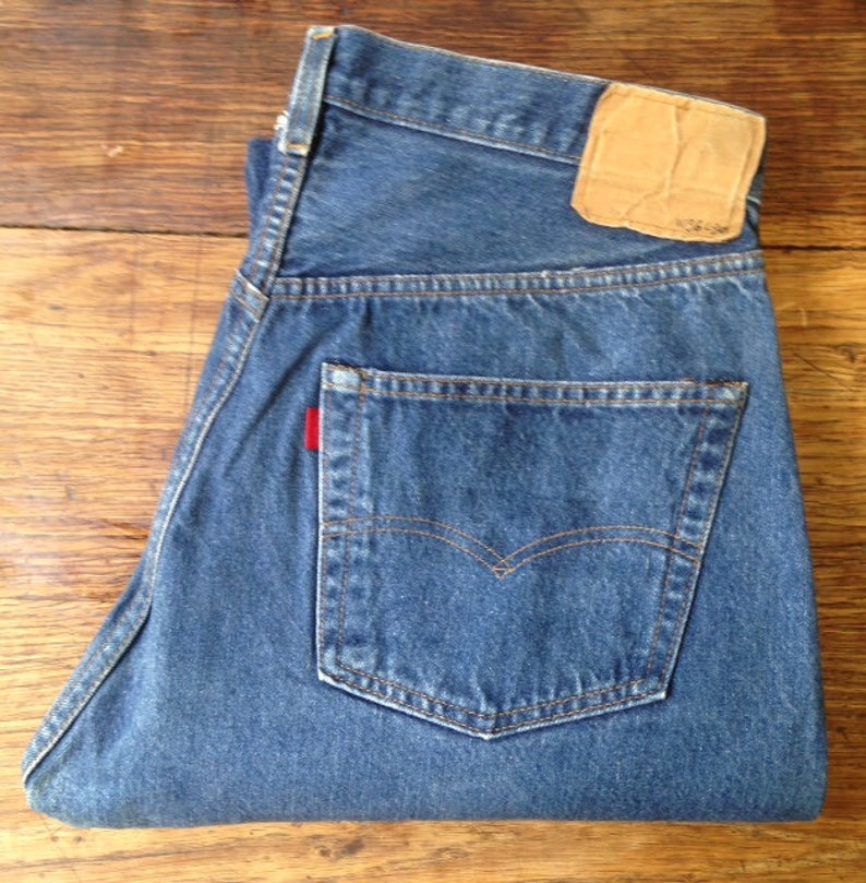 May include: A pair of medium-wash blue denim jeans featuring a tan leather patch on the back waistband, a single rear pocket with orange contrast stitching, and a small red fabric tab attached to the side of the pocket seam.