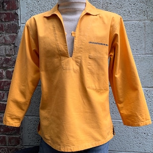 May include: Mustard yellow long-sleeve shirt with a notched collar and a small pocket on the chest. The shirt has a V-neck opening and a button closure. The text "LES SAUVETEURS EN MER" is printed on the chest.
