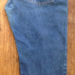May include: A pair of medium-wash blue denim jeans featuring a high-rise waist, a tan leather-like patch on the back waistband, a single back pocket with a small red tab, and a straight-leg cut with a folded cuff at the bottom hem.