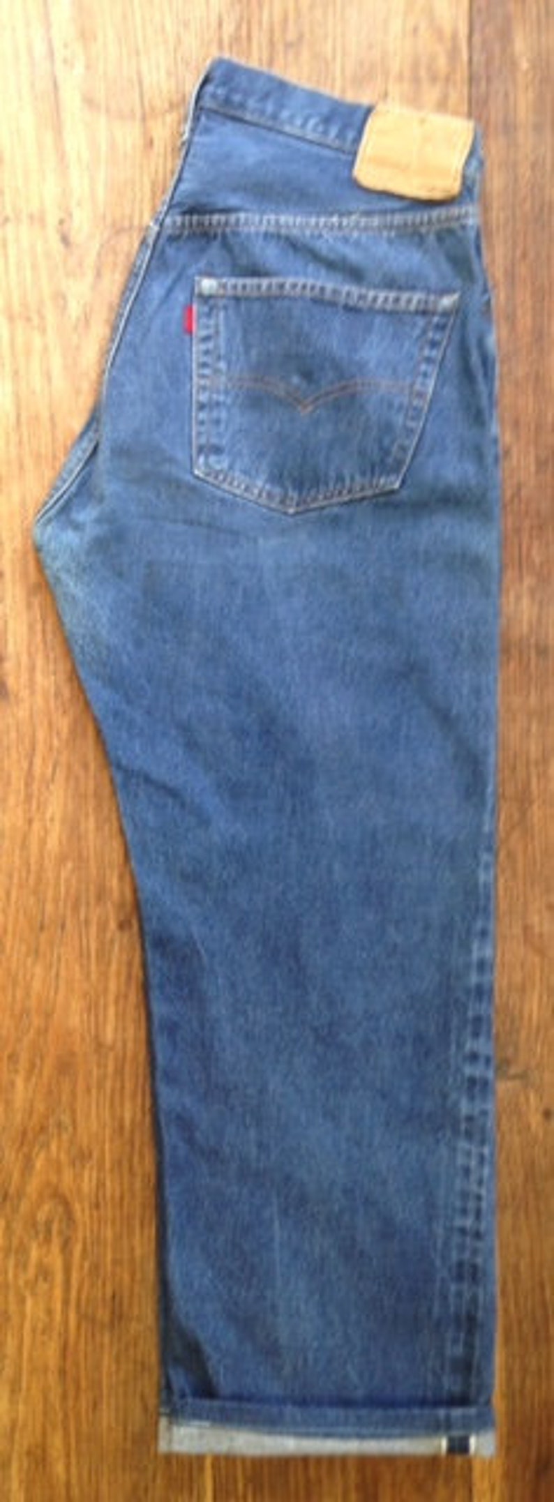 May include: A pair of medium-wash blue denim jeans featuring a high-rise waist, a tan leather-like patch on the back waistband, a single back pocket with a small red tab, and a straight-leg cut with a folded cuff at the bottom hem.