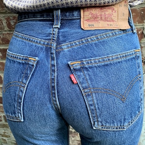 May include: Close-up of the back of blue denim jeans with a red tag on the back pocket. The jeans have a classic five-pocket design, with orange stitching and a leather patch. The waist size is W26 L32.