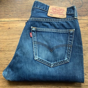 May include: A pair of blue denim jeans with a brown leather Levi's patch on the back pocket. The patch has the text "Levi Strauss & Co." and "Quality Clothing" printed on it. The jeans have a red Levi's tab on the back right pocket. The jeans are folded in half and the back pockets are visible.