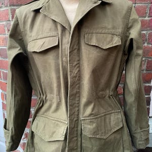 Peut inclure: Veste de style militaire vert olive avec quatre poches avant et une taille ajustable par cordon. La veste a un col et des poignets boutonnés. Un col roulé crème est visible en dessous.