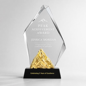 May include: A clear, angular glass award featuring a gold-colored mountain sculpture at the base. The glass is etched with a mountain icon, the text PEAK ACHIEVEMENT AWARD, JESSICA MORGAN, In Appreciation of Your Leadership & Dedication, and a black base.