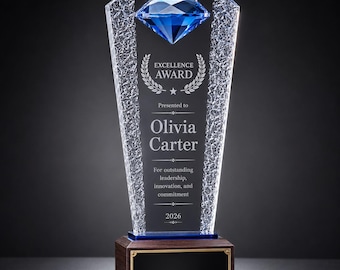 Pode incluir: Um troféu de vidro transparente com um diamante azul facetado no topo, painéis laterais texturizados e uma base de madeira escura com uma placa preta em branco. O vidro está gravado com o texto EXCELLENCE AWARD, Presented to Olivia Carter, For outstanding leadership, innovation, and commitment, e 2026.