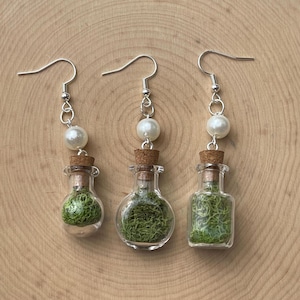 May include: Three pairs of earrings featuring small glass bottles filled with green moss, topped with cork stoppers and a white pearl charm. The earrings have silver hooks.