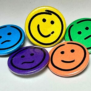May include: Five round buttons with different coloured backgrounds and black outlines. Each button features a hand-drawn smiley face with different expressions: happy, sad, neutral, confused, and surprised.