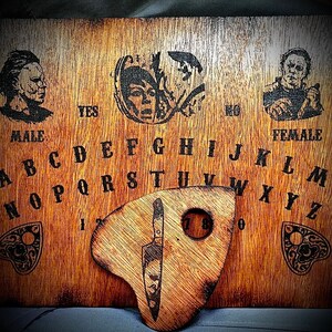 May include: A wooden Ouija board with a planchette in the shape of a knife. The board has a black and white alphabet, numbers 1-9, and the words "yes", "no", "female", and "male".