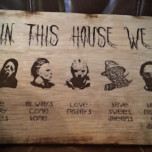 Puede incluir: Un letrero de madera con el texto "IN THIS HOUSE WE" e imágenes de personajes de películas de terror. El texto debajo de las imágenes dice "Like scary movies, always come home, love Fridays, have sweet dreams, have friends for dinner."