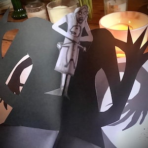May include: A black paper cutout of a monster with long arms and sharp claws. The monster is holding a white paper cutout of a woman with a child in her arms. The woman is looking up in fear.