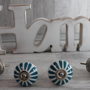 Könnte beinhalten: Vier weiße Keramik-Schrankknöpfe mit einem blaugrünen und weißen Blumenmuster. Die Knöpfe haben einen silbernen Metallakzent in der Mitte.