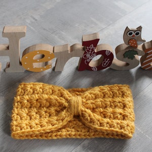 Könnte beinhalten: Ein senfgelbes Häkel-Stirnband mit einer Schleife in der Mitte. Das Stirnband befindet sich vor Holzbuchstaben, die "Herbst" buchstabieren, das deutsche Wort für "Herbst".