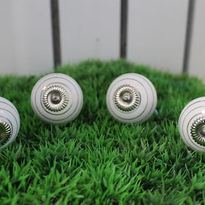 Könnte beinhalten: Vier weiße Keramik-Schrankknöpfe mit silbernem Mittelteil und einem dünnen goldenen Rand. Die Knöpfe liegen auf einem Bett aus grünem Kunstgras.