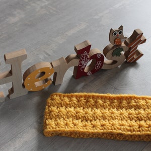 Könnte beinhalten: Ein gelbes Häkelband mit einem strukturierten Muster. Das Stirnband liegt auf einer Holzoberfläche mit dem Wort "Herbst" in Holzbuchstaben.