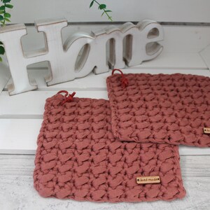 Könnte beinhalten: Zwei gehäkelte Topflappen in einem staubigen Rosaton mit einem Holzschild, auf dem "hand made" steht. Die Topflappen liegen auf einer weißen Holzoberfläche mit einem weißen Holzschild, auf dem "Home" steht.