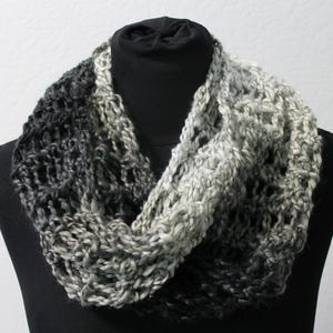 Könnte beinhalten: Ein handgestrickter Loop-Schal in Grau- und Weißtönen. Der Schal wird auf einer schwarzen Schaufensterpuppe präsentiert, die seine offene, strukturierte Webart zeigt. Der Farbverlauf geht von Dunkelgrau zu Hellgrau über.