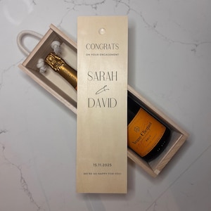 May include: A wooden champagne gift box containing a bottle of Veuve Clicquot Brut champagne. The box is personalised with the names Sarah & David, the date 15.11.2025, and the text "Congrats on your engagement".