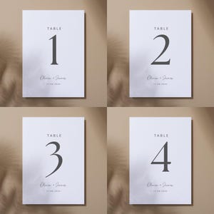May include: Four white table number cards with black text. Each card displays a large number from one to four, the word "TABLE", the names "Olivia + James", and the date "11.06.2024". The cards are on a neutral background.
