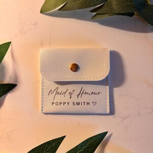 May include: An off-white pouch with a flap secured by a gold-tone stud. The text "Maid of Honour" is printed in script, with the name "Poppy Smith" and a heart symbol below. The pouch is likely a wedding-related gift.