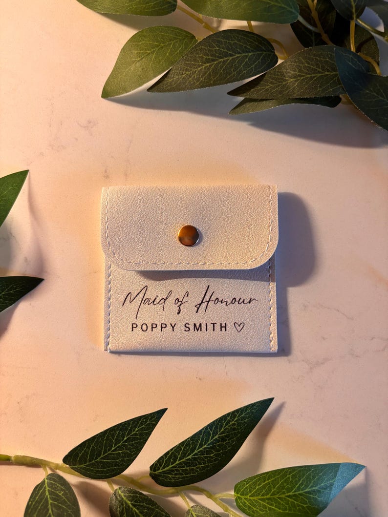 May include: An off-white pouch with a flap secured by a gold-tone stud. The text "Maid of Honour" is printed in script, with the name "Poppy Smith" and a heart symbol below. The pouch is likely a wedding-related gift.